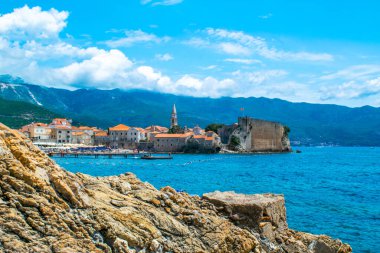 Eski şehir duvarları kalesinin manzarası. Turuncu çatıları olan eski Budva kasabasının mimarisi. Ön planda taş var. Güzel mavi yaz güneşli dağ manzarası. Karadağ.