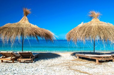 Saman plajı şemsiyeleri ve temiz kum ve çakıl taşı plajındaki rahat güneş şezlongları. Himare. Arnavutluk. İyon Denizi. Tatil deniz konsepti. Yaz güneşli deniz manzarası.