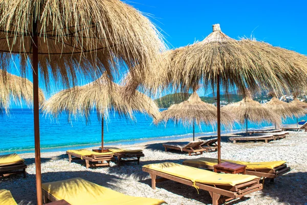 Saman plajı şemsiyeleri ve temiz kum ve çakıl taşı plajındaki rahat güneş şezlongları. Himare. Arnavutluk. İyon Denizi. Tatil deniz konsepti. Yaz güneşli deniz manzarası.