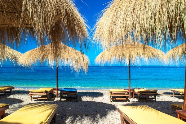 Saman plajı şemsiyeleri ve temiz kum ve çakıl taşı plajındaki rahat güneş şezlongları. Himare. Arnavutluk. İyon Denizi. Tatil deniz konsepti. Yaz güneşli deniz manzarası.