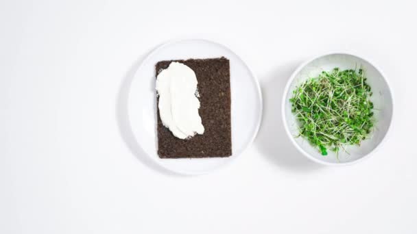 4Kマイクログリーンの緑の芽を持つボウルの近くに 黒いパンのプレートが表示されます サンドイッチはクリームチーズで広がり 緑の芽がトッピングされています 白地だ ストップモーションアニメーション — ストック動画