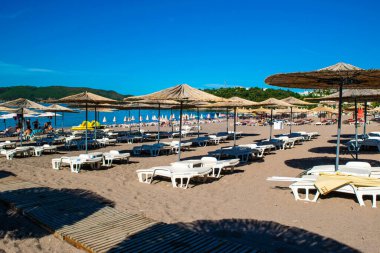Becici, Karadağ - 02 Temmuz 2021: Yaz sabahları halk kumsalında dinlenen insanlar, dinlenme yerleri ve şemsiyeler. Sakin deniz. Mavi gökyüzü. Budva Riviera. Deniz kenarı tatil köyü sezonu.