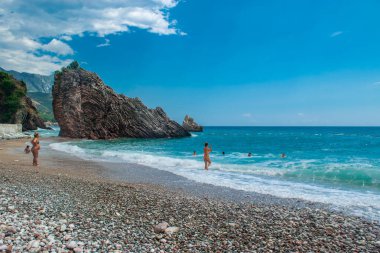 Rafailovici, Karadağ - 05 Temmuz 2021: Çakıl taşlı plajda güzel kayalarla dinlenen insanlar. Adriyatik Denizi. Yaz güneşli bir gün. Deniz kenarı tatil köyü sezonu.