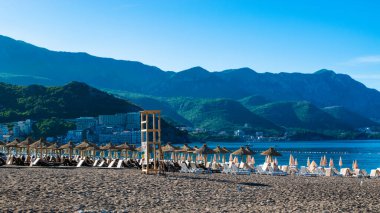 Sıralar dolusu plaj şemsiyesi ve güneş şezlongları sabah güneşinde güzel kıyı dağlarının zeminine karşı. Sakin deniz ve mavi gökyüzü. Budva Riviera. Deniz kıyısındaki tatil köyü sezonu. Becici, Karadağ.