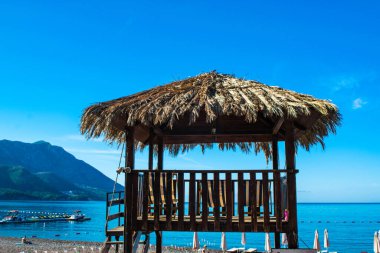Halka açık kumsalda sazdan gölgelikli bir platform ve güneşli salonlar. Mavi gökyüzünün arka planı ve sakin Adriyatik Denizi. Güneşli bir sabah. Budva Riviera. Deniz kıyısındaki tatil köyü sezonu. Becici, Karadağ.