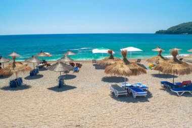 Saman plajı şemsiyeleri ve temiz kum ve çakıl taşı plajındaki rahat güneş şezlongları. Himare. Arnavutluk. İyon Denizi. Tatil deniz konsepti. Yaz güneşli deniz manzarası.