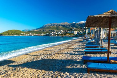 Güzel, temiz kum manzarası ve kumsal şemsiyeli çakıl taşı plajı ve dağ arkasındakiler. Himare. Arnavutluk. İyon Denizi. Tatil deniz konsepti. Yaz güneşli mavi deniz manzarası