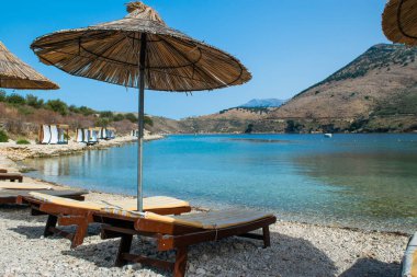 Sun loungers, umbrellas and comfortable beds canopy on the Porto Palermo beach near old fortress Ali Pasha Tepelena. Ionian Sea. Albania. Blue summer landscape.