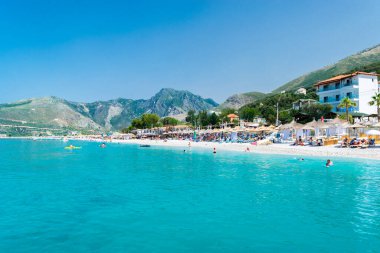 Borsh Beach, Albania - July 14, 202: Vacationers on pebble beach with clear blue water at foot of mountains.  Ionian Sea. Summer landscape. Concept of summer holidays and relaxation.