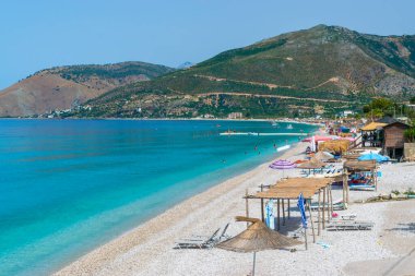 Vacationers on pebble Borsh beach with clear blue water at foot of mountains. Albania Ionian Sea. Summer landscape. Concept of summer holidays and relaxation.