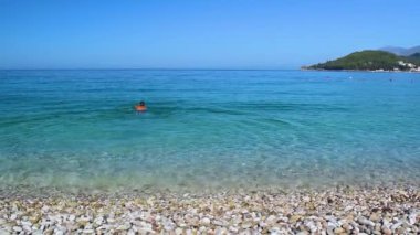 Young man with swarthy skin and black hair is swimming in the sea. Beautiful clean pebble Potam beach with clear blue water. Himare. Albania. Ionian Sea.  Summer sunny landscape.