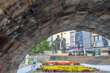 ÜSKÜP, NORTH MACEDONYA-AĞUSTOS 28. 2018: Bir erkek bisikletçi, Taş Köprü kemerinin altından bakıldığında, iki çiçek yatağı arasındaki yolu geçer.