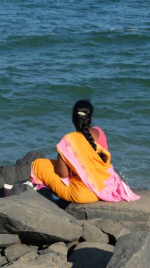 PONDICHERRY / INDIA-FEBRUARY 23 2018: Sari giymiş Hintli kadın bir kayanın üzerinde oturuyor ve denize bakıyor.