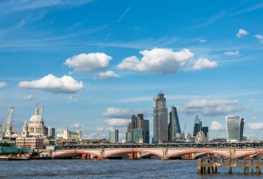 Blackfriars köprüsü, açık bir yaz öğleden sonra Thames nehrinden geçer, St. Pauls katedrali ve diğer ikonik şehir binalarıyla birlikte, mavi gökyüzüne ve tüylü beyaz bulutlara karşı..