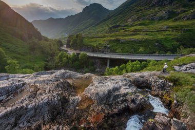 Glencoe,Scotland,UK-July 18th 2022: A lone figure surveys the main road of Glencoe,in the Scottish Highlands,as it winds it's way into the distant valley,close to evening sunset, during mid summer.