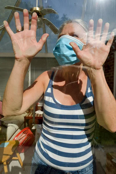 A housewife wearing a protective face mask during Corona virus lock down pressing her hands against the glass feeling isolated,desperate to be outside with loved ones and uncertain about the future.