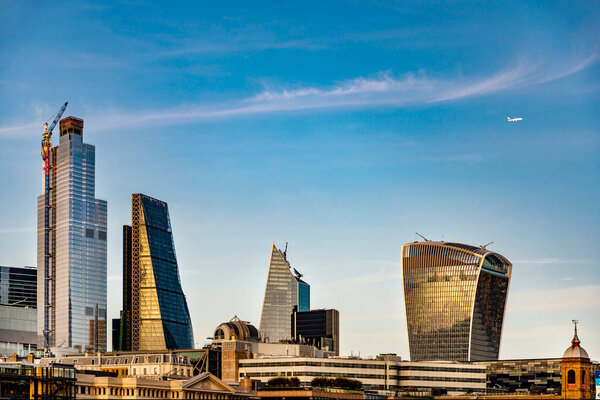 A distant A380 passenger jet leaving Heathrow,moves across clear blue sky, behind familiar modern City of London buildings,reflecting light from the setting sun over the River Thames.
