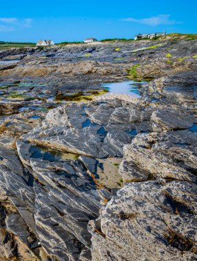Kuzey Cornwall, İngiltere, İngiltere-20 Temmuz 2021: Uzak tatilciler, güneşli yaz mevsiminin tadını çıkararak Cornish kulübelerinin yakınında yürürler ve ön planda, dalgalı granit kayalar yüksek gelgiti bekler..