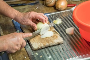 Yaşlı kadın mutfakta soğan doğruyor lavabo ve kırmızı plastik kasenin yanında tahta tahta ve bıçakla.