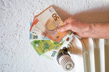 Almanya 'da bir apartmanda ısıtma termostatı ve radyatör önünde elinde banknotlar tutan yaşlı bir vatandaş.