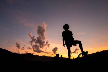 Dağların gölgeleriyle çevrili bir kütüğün üzerindeki genç bir çocuğun silueti. Akşamları yere düşüyor. Egzersiz sonrası dinlenme hissi..