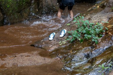Küçük bir şelalenin yanındaki kayaya yerleştirilmiş seçici beyaz sandaletler. Bir çocuk köyün yakınındaki ormandaki bir su kaynağında koşup oynuyor.