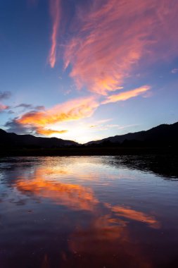 Manzara silueti Dağlar, akşam gökyüzünde gün batımı ve sonra bulutlara vuran ışık turuncu renktedir. Nehirde rahatlamış hisseden bir yansıma var. Metin için yer var..