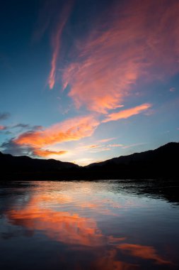 Manzara silueti Dağlar, akşam gökyüzünde gün batımı ve sonra bulutlara vuran ışık turuncu renktedir. Nehirde rahatlamış hisseden bir yansıma var. Metin için yer var..