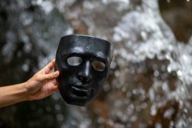 Onu tutan iş adamının elindeki siyah maskenin odak noktasını seçin. Doğa şelalesi arka planı. Kavram. Maskeni çıkar, kimliğini açıkla ve doğaya geri dön. Karanlık yanını çıkar..