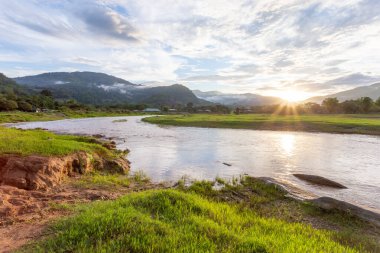 Dağ manzarası, gün batımı, sis, dağdaki yağmurdan sonraki bulutlar, berrak gökyüzü, nehir, açık yeşil çimenler, arka planda serin ve ferahlatıcı bir his verir.