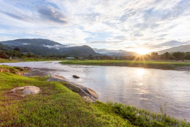 Dağ manzarası, gün batımı, sis, dağdaki yağmurdan sonraki bulutlar, berrak gökyüzü, nehir, açık yeşil çimenler, arka planda serin ve ferahlatıcı bir his verir.