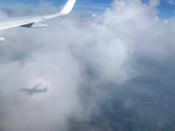 Airplane Window view on white wing and white clouds and infinite horizon by clear sunny weather, with a visible airplane shadow rounded by rainbow circle