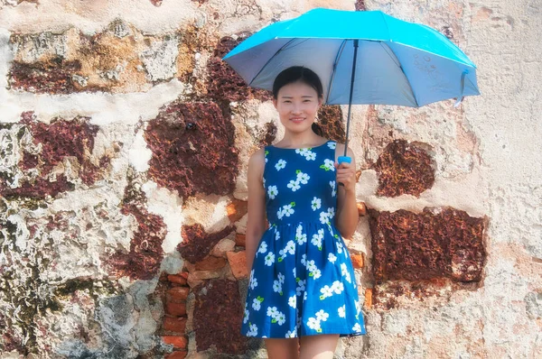 在阳光灿烂的一天 一位快乐的中国女人拿着一把雨伞 穿着花衣 站在风风雨无阻的砖墙边 — 图库照片