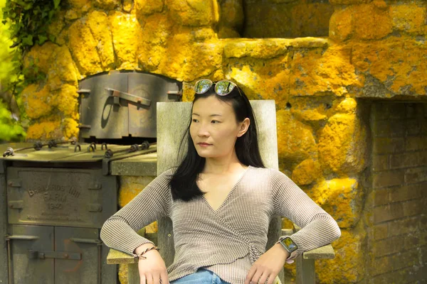A chinese woman sitting in a chair in front of an outside fireplace looking away from the camera.