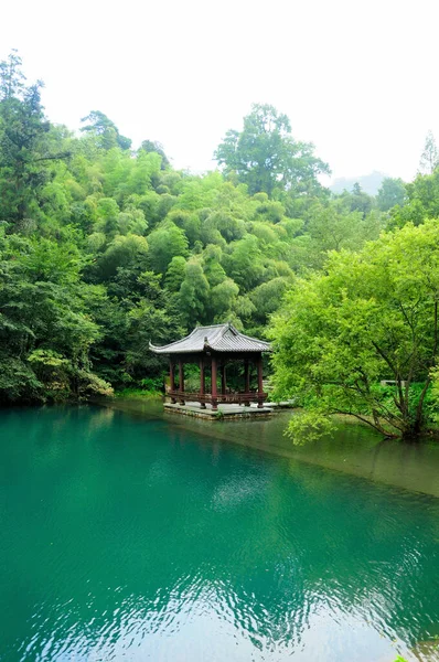 A chinese gazebo sitting on an azure colored water against a lush grove of trees.