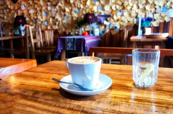 A white coffee cup with cappuccino and a glass of water on a wooden table within a coffee shop inside of Fenghuang Ancient town, Hunan province China.
