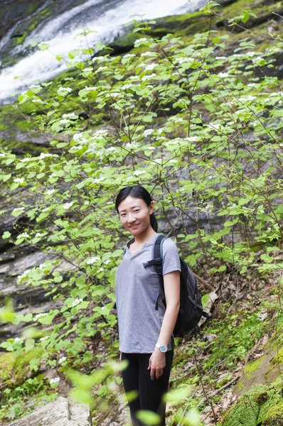 A smiling chinese woman surrounded by hobblebush vibrunum with a blurred waterfall background outside in falls village connecticut new england.