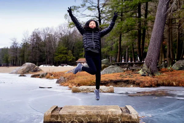 A chinese woman jumping and flashing peace signs on a cememt block at Burr Pond State Park in wintertime in Torrington Connecticut.