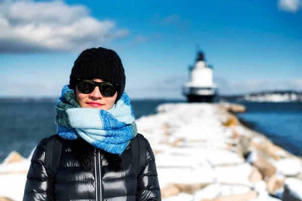 A smiling chinese woman bundled up from the cold on the jetty leading to the Spring Point Ledge Lighthouse in South Portland Maine on a blue sky winter day.