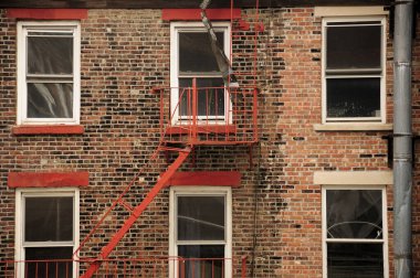 The exterior of a red brick building as a metal fire escape is located near one of the windows in New York City.  
