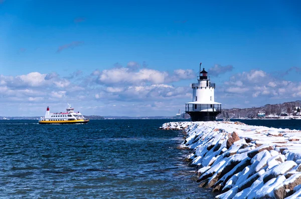 A passenger ferry traveling near the snow covered spring point ledge lighthouse in portland maine on a blue sky winter day.