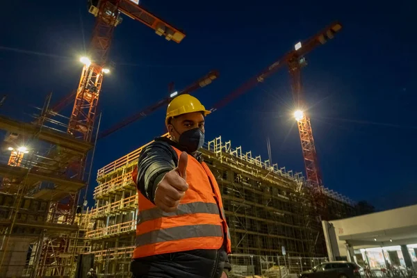 Construction worker outdoors at night wearing a safety vest, a medical mask and a protective hardhat and showing thumbs up gesture