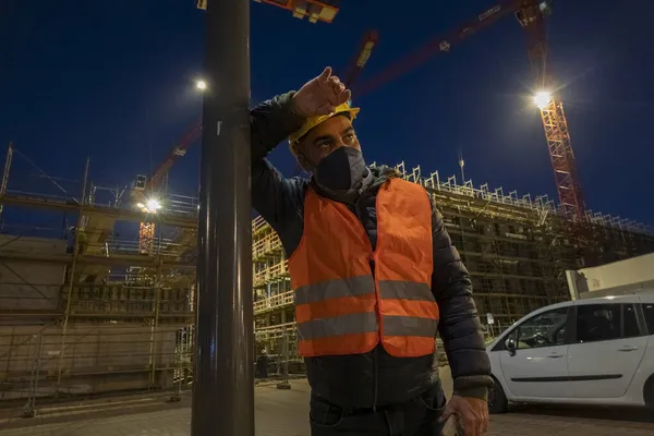 Tired and pensive construction worker outdoors at night wearing a safety vest, a medical mask and a protective hardhat leaning against a light pole