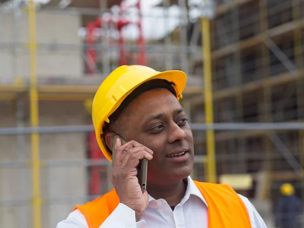 印度或索马里面带微笑的工程师 身穿安全夹克 头戴安全帽 在建筑工地用手机交谈 免版税图库图片