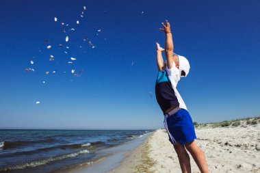 Küçük çocuk sahilde deniz kabuklarıyla oynuyor. Mutlu çocuk yazın, denizin ve tatilin tadını çıkarıyor.