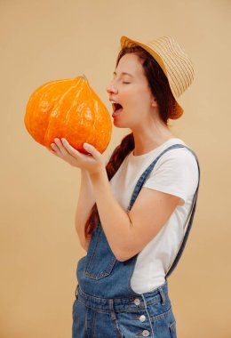 Çiftçi kadın bej bir arka planda olgun bir balkabağını ısırıyor..