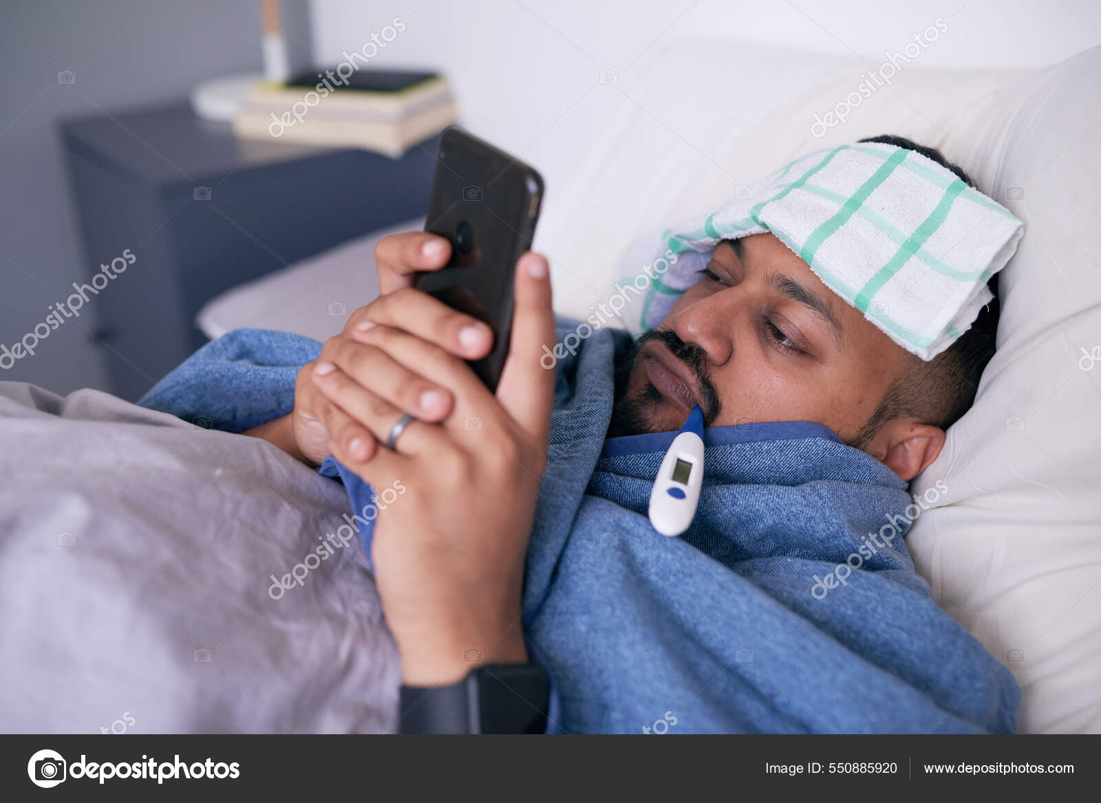 A sick man in bed with a thermometer looks at his mobile device Stock Photo  by ©MeekoMedia 550885920