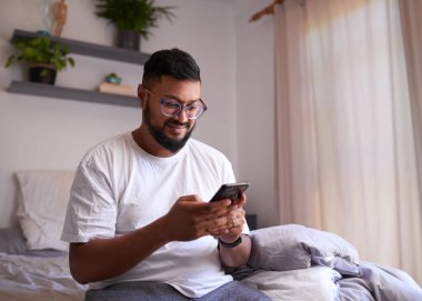 Bir adam sabah yatak odasında cep telefonuna gülümsüyor.