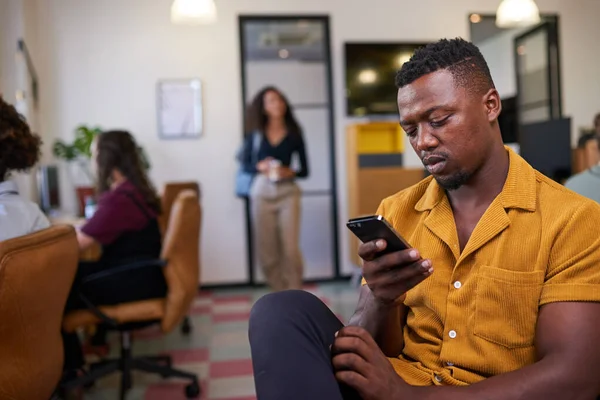 Um homem rola em seu celular olhando entediado no trabalho no escritório ocupado — Fotografia de Stock