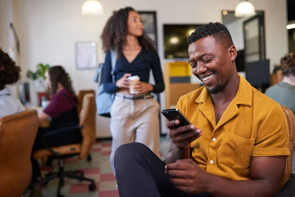 Um empresário negro olha para seu telefone em um escritório diversificado ocupado — Fotografia de Stock